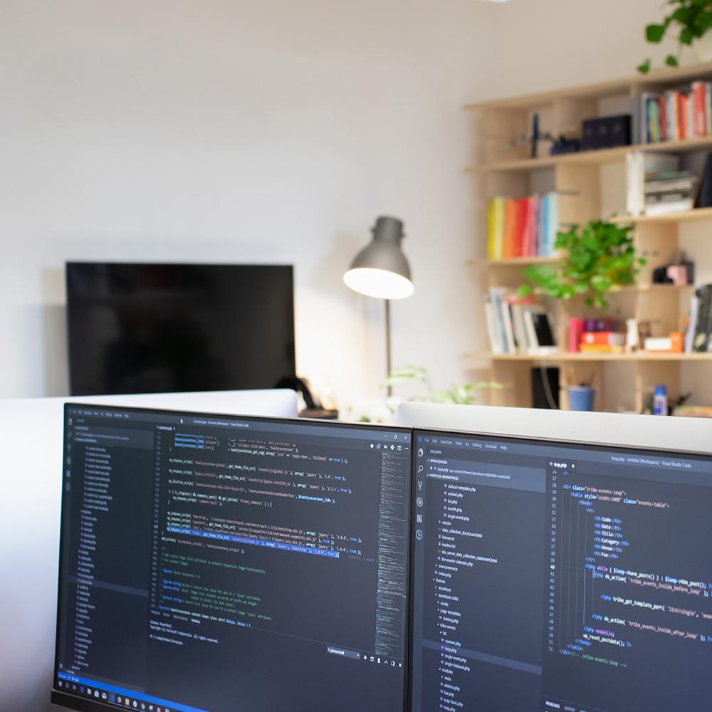A modern, bright developer workspace with dual monitors displaying code, surrounded by plants, bookshelves and warm lighting, with colourful abstract wall decorations.